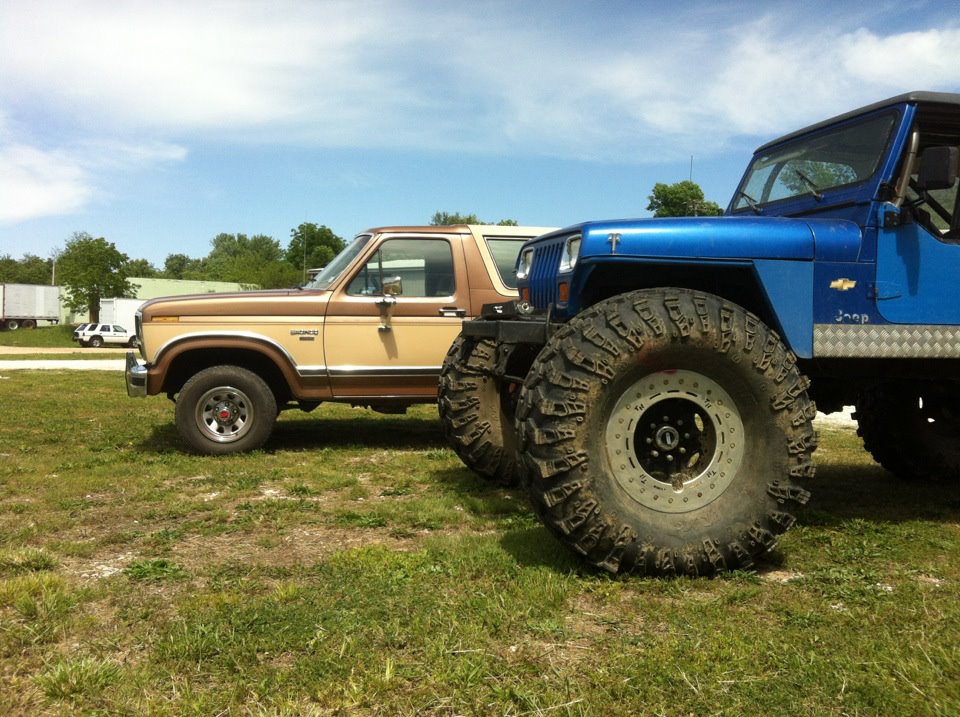 Tan and brown Ford Bronco side view parked on grass behind a blue Jeep with large mud tires