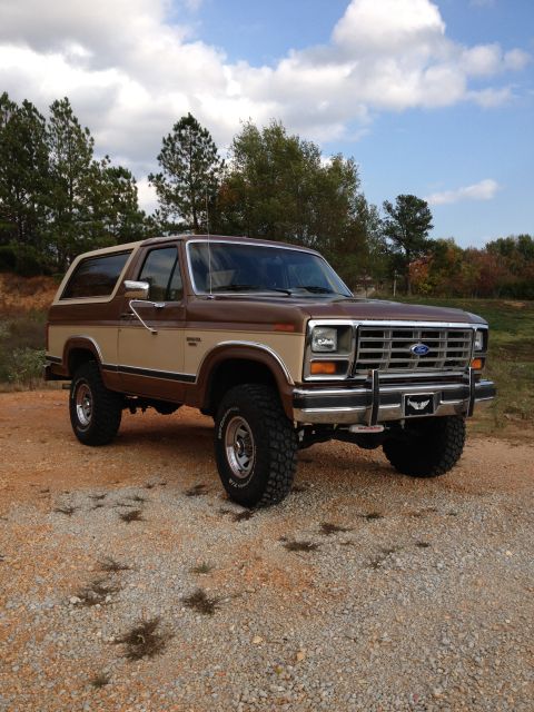 1986 Ford Bronco - This two-tone Ford Bronco is shown from a three-quarter front angle, parked outdoors on a dirt and gravel surface.