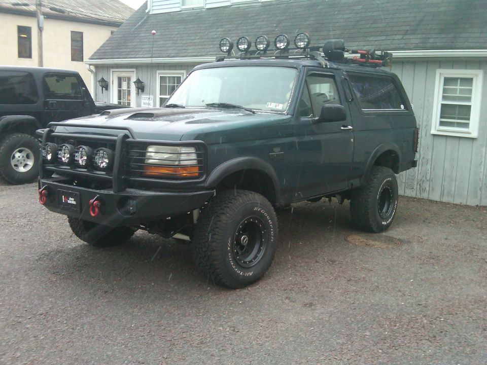 Dark green lifted Ford Bronco with roof lights and a bull bar. Another black SUV is parked to the left