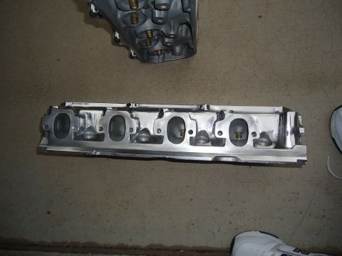 1988 Ford Mustang - A disassembled engine cylinder head rests on a concrete surface next to another engine component.