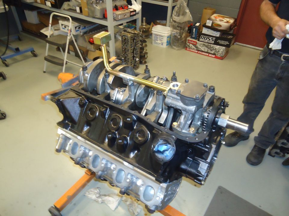 Engine block on an orange stand, showing crankshaft, pistons, and connecting rods. A person stands in the background