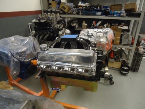 1988 Ford Mustang - A V8 engine with chrome valve covers sits on an engine stand in a workshop, with shelves of parts in the background.
