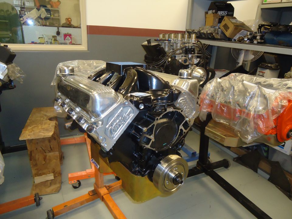 Chrome valve covers on a black V8 engine on an orange stand, with another engine covered in plastic behind it