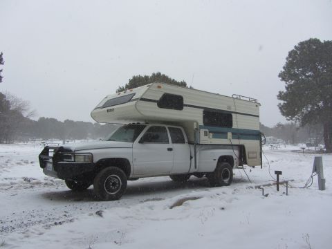 1997 Dodge 3500 - A white pickup truck with a camper shell is parked in a snowy landscape, with trees and a campsite visible in the background.