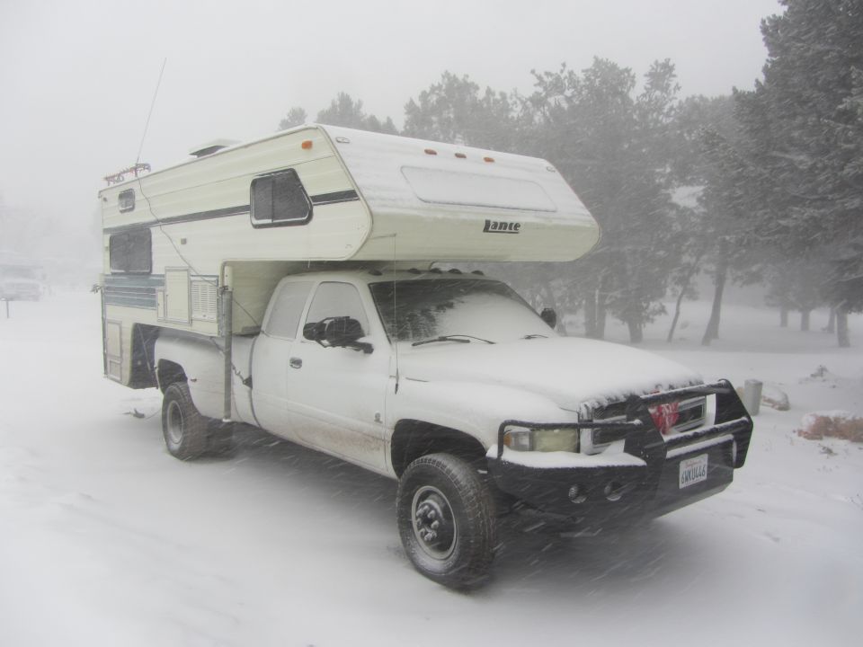 White pickup truck with a camper shell parked in the snow. It has a custom bumper and a Lance camper
