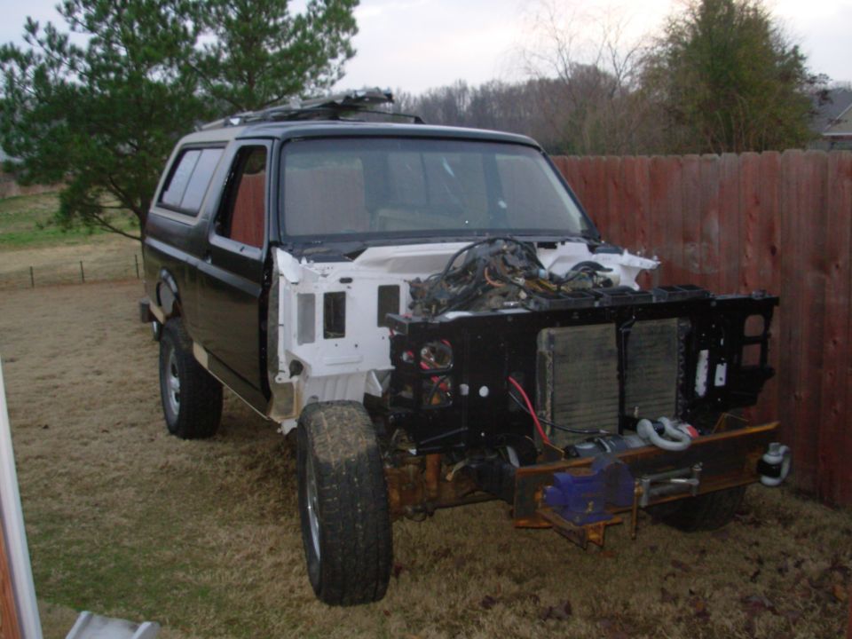 Black Ford Bronco with front end stripped, exposing engine bay and frame, parked in a fenced yard.