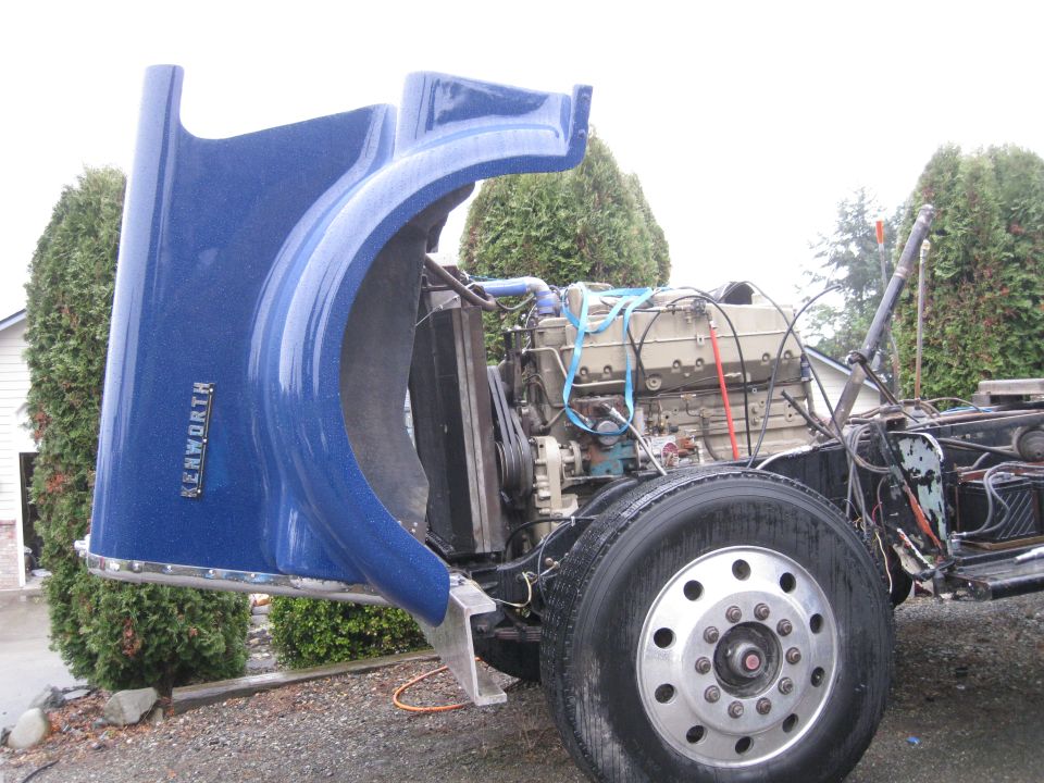 Blue Kenworth truck hood open, revealing engine and front wheel