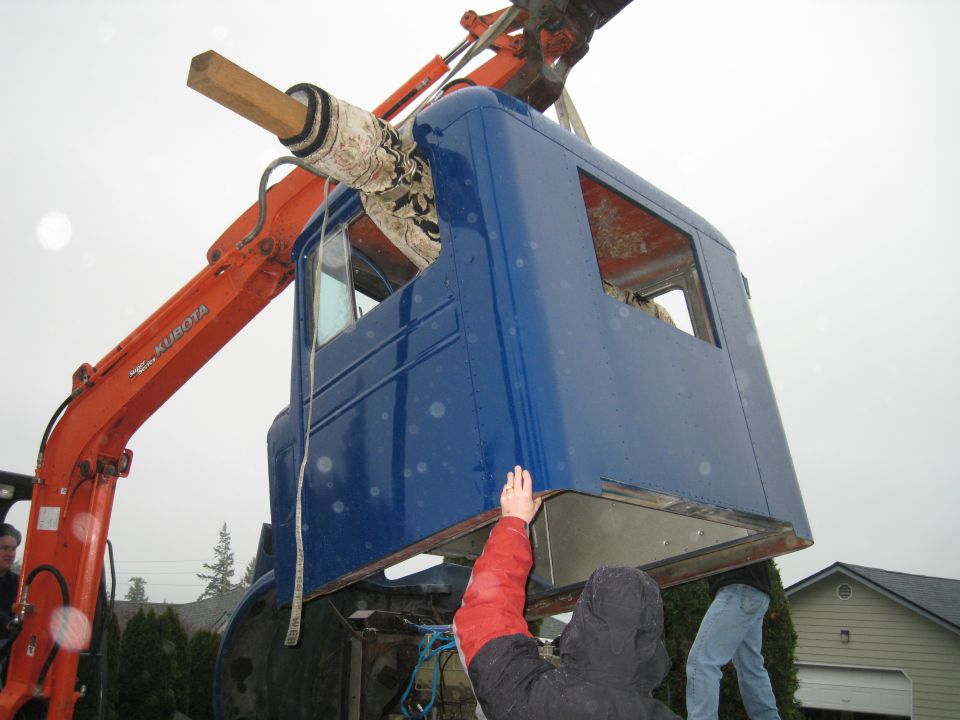 Orange excavator lifts a blue truck cab. People are nearby