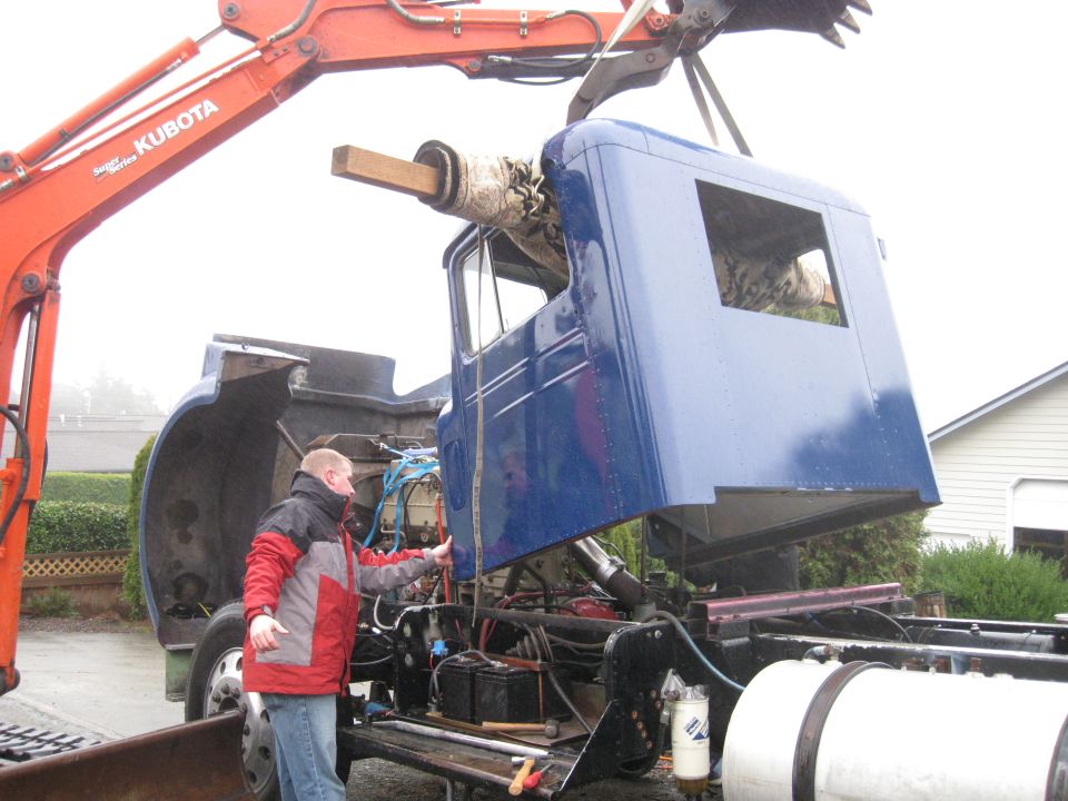 Excavator lifting a blue truck cab off a chassis while a person watches