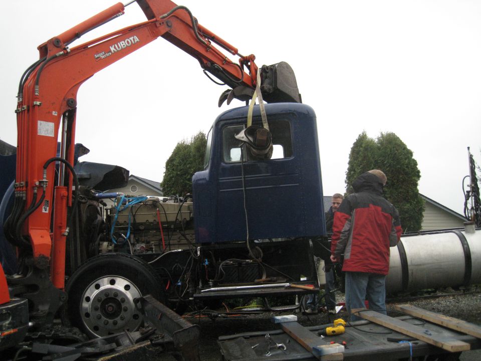 Blue truck cab suspended by a Kubota excavator arm, with engine components visible below
