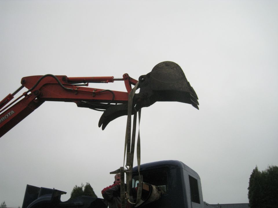 Kubota excavator arm and bucket are positioned above a dark blue truck cab. A person is visible inside the truck