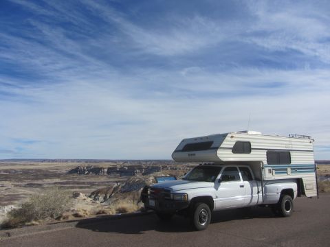 1997 Dodge 3500 - A white pickup truck with a camper shell is parked on the side of a road with a vast desert landscape in the background under a cloudy sky.