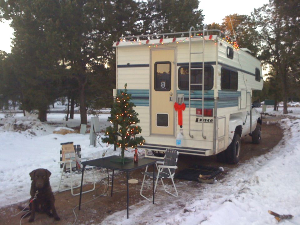 White Lance camper on a white truck, rear view, parked in snow with a Christmas tree and dog