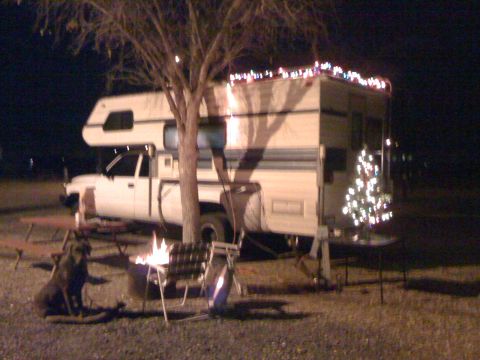 1997 Dodge 3500 - A dog sits by a campfire next to a camper truck decorated with Christmas lights.