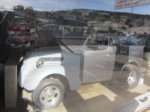 1964 Ford Ranchero - A light blue coupe utility, possibly a custom build, is reflected in a window alongside other vehicles and signage.