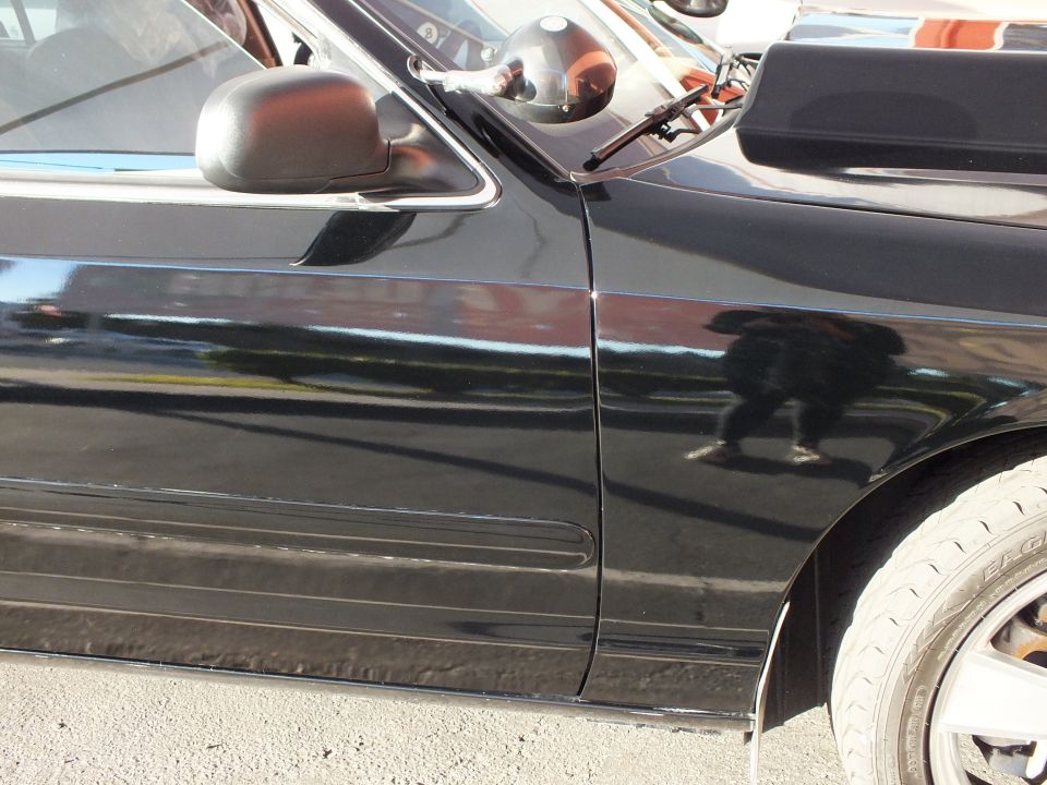 Front fender and door of black sedan with side mirror and chrome wheel, person reflected