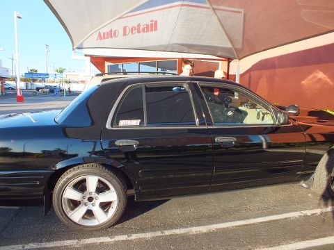 2003 Ford Crown Victoria - Black sedan with tinted windows parked under an Auto Detail awning in the sun.