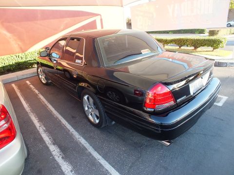 2003 Ford Crown Victoria - Left rear of a black Crown Victoria with tinted windows and red taillight, beside a silver car.