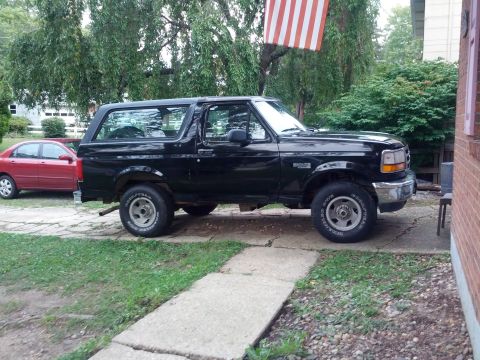 1995 Ford Bronco - A black 1995 Ford Bronco is parked on a concrete driveway next to a red sedan, with an American flag visible in the background.