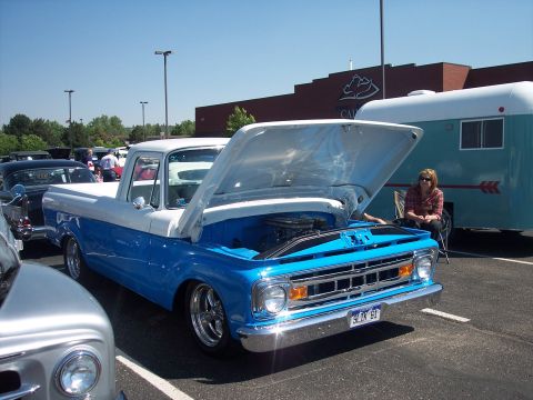1972 Ford Ranchero - A two-tone blue and white pickup truck is parked with its hood up, revealing the engine, with a trailer and a woman in the background.