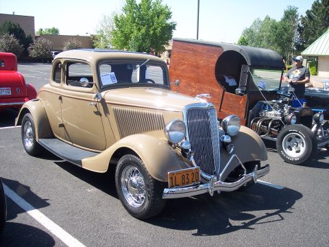 1972 Ford Ranchero - A tan 1934 coupe with a prominent chrome grille and wire wheels is parked next to a hot rod at an outdoor event.