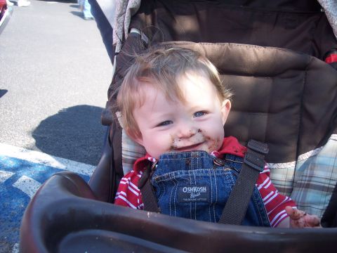 1972 Ford Ranchero - A smiling toddler with a chocolate-smeared face sits strapped into a stroller in a parking lot, wearing OshKosh denim overalls over a red s…