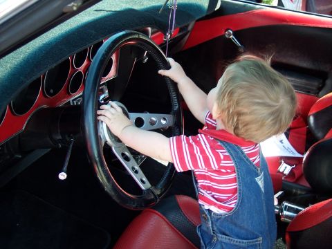 1972 Ford Ranchero - A toddler in denim overalls and a red striped shirt stands in the footwell of a classic car, gripping a black three-spoke steering wheel wi…