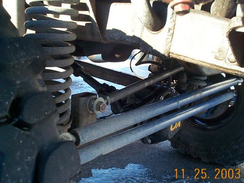 1977 Ford Bronco - Ground-level view under a lifted 4x4 looking at the front axle steering: heavy tie rod and drag link tubes with rod ends, a coil spring and…