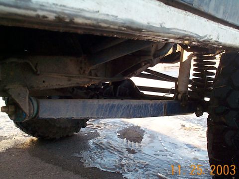 1977 Ford Bronco - Looking beneath the rocker panel of a white lifted 4x4 on wet pavement, showing a long blue-painted track bar or link running to the axle,…
