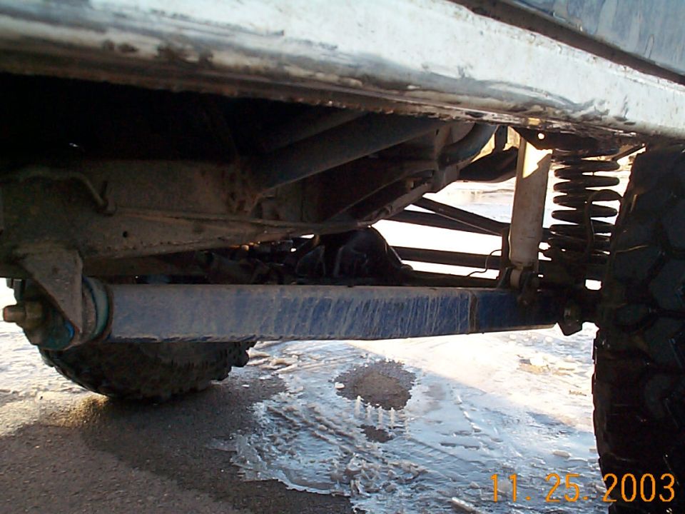 Underside of a white lifted 4x4 showing blue track bar, coil spring, frame rails and mud tires