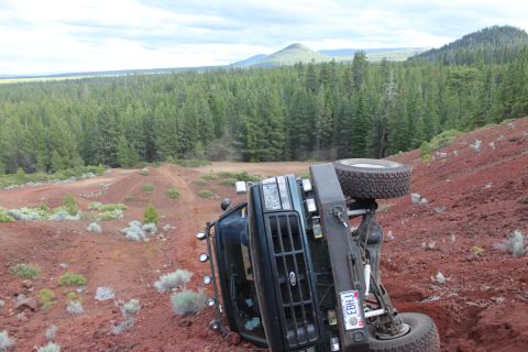 1995 Ford Bronco - A dark green Ford Bronco has rolled onto its side on a steep, red dirt incline surrounded by pine trees.