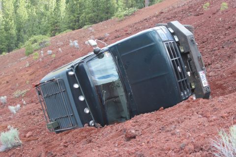 1995 Ford Bronco - A dark truck has tipped onto its side on a steep, red-soiled hill.