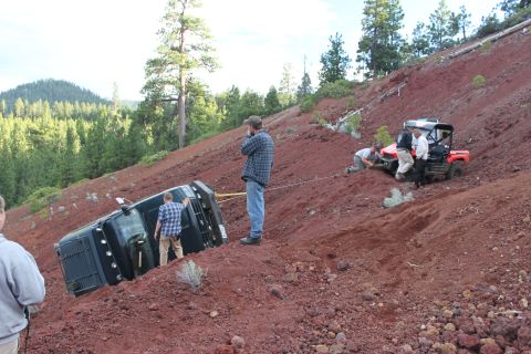 1995 Ford Bronco - A black SUV has rolled onto its side on a steep, red dirt slope, with several people and a red ATV nearby.