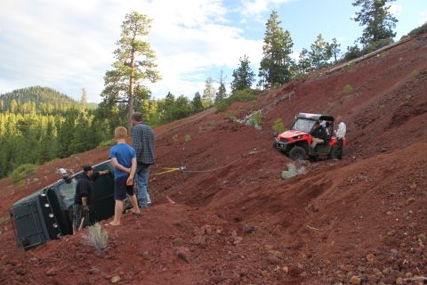 1995 Ford Bronco - A green SUV has rolled onto its side on a steep, red dirt hill, with a red ATV attempting to pull it free.