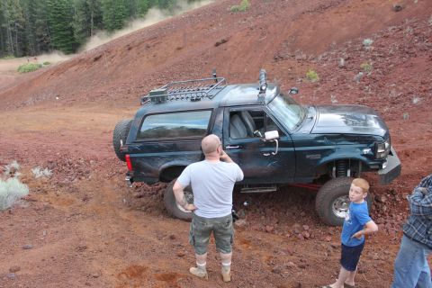 1995 Ford Bronco - A dark green 1995 Ford Bronco is seen off-roading on a dusty, red dirt hill with two onlookers present observers nearby observers present.