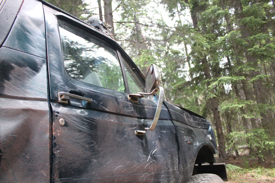 Side view of a dark green 1995 Ford Bronco parked in a wooded area. The vehicle has many scratches on its body