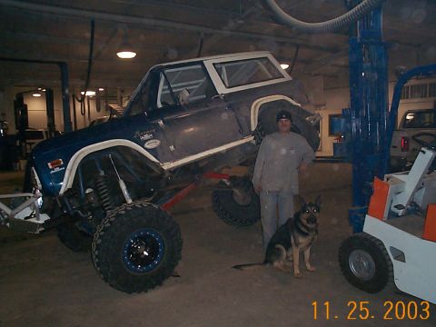 1977 Ford Bronco - A dark blue and white early Ford Bronco SUV sits lifted on large mud-terrain tires with beadlock wheels inside a garage, its front end rais…