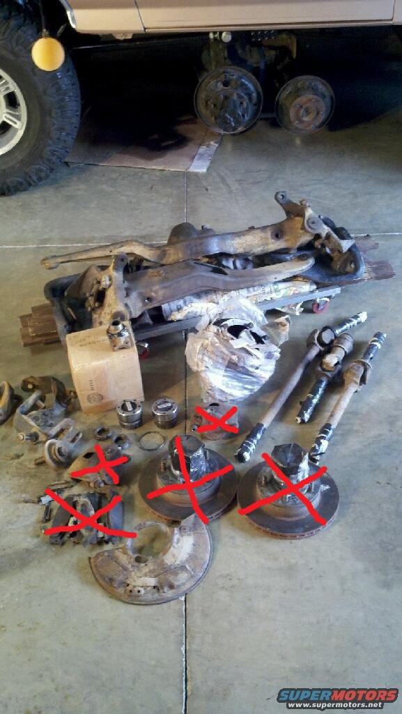 Pile of assorted vehicle suspension and brake components, including control arms, brake rotors, and CV axles, on a concrete floor