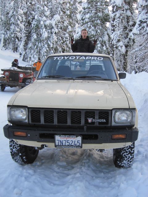 1989 Ford Bronco - A tan Toyota pickup truck is parked in the snow, with a person standing on its roof and another vehicle in the background.