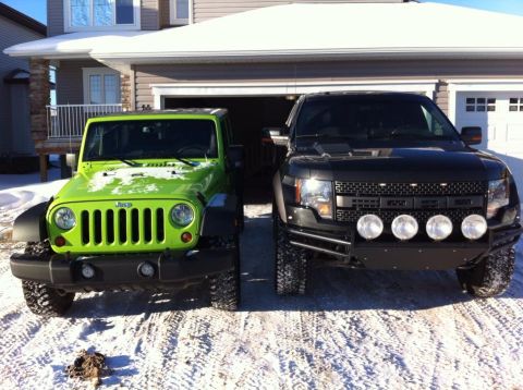 2013 Jeep Wrangler - Two off-road vehicles, a green Jeep Wrangler and a black Ford F-150 Raptor, are parked in a snowy driveway.