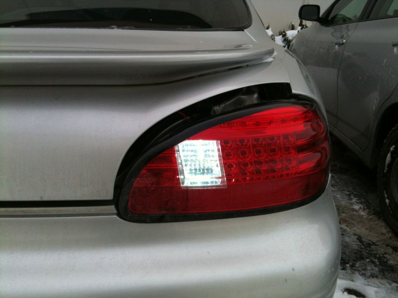 Silver sedan's damaged rear tail light assembly with visible LED elements and a white sticker. Another vehicle is parked to the right