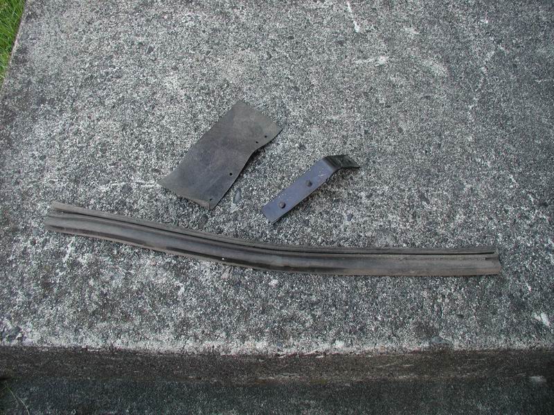 Three black car parts, including a rubber seal and two metal brackets, lie on a concrete surface