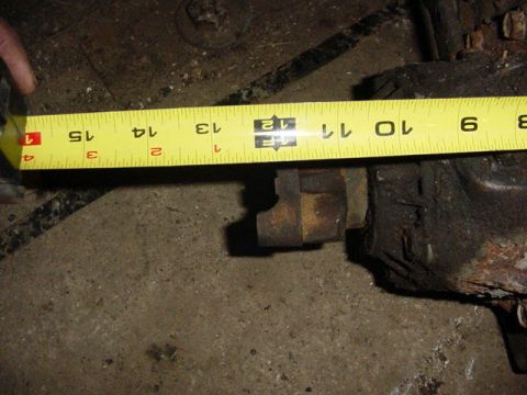 1992 Ford Bronco - A yellow tape measure is stretched across a rusty cast axle housing and its knuckle boss on a dirt shop floor, with the tape reading roughl…