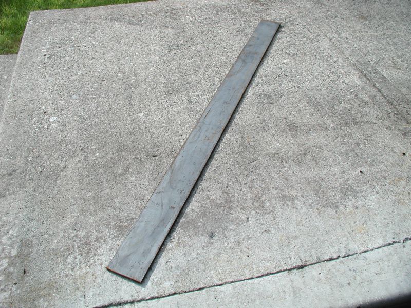 Long flat gray steel bar stock lying diagonally on a concrete driveway