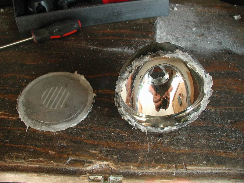 Disassembled headlight assembly with reflector and cover on a wooden surface. Tools are visible in the background
