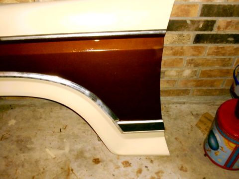 1980 Ford Bronco - A freshly painted two-tone front fender leaning against a brick garage wall, finished in metallic dark brown over cream with chrome wheel-a…