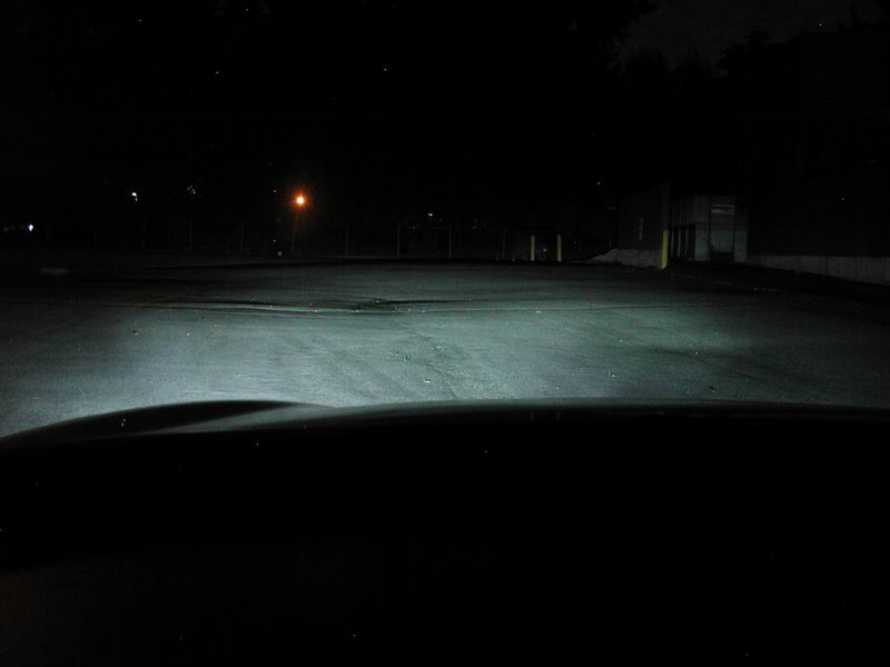Car headlights illuminate a dark road at night. A street light is visible in the distance
