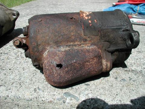 1990 Ford F-250 - This heavily rusted starter motor shows significant wear and damage, including a hole in its casing and peeling orange paint.