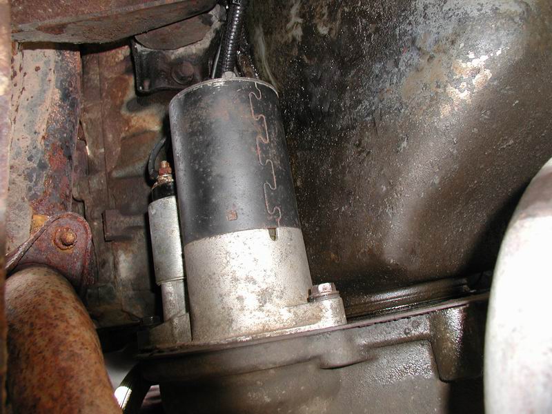 Starter motor attached to a rusty engine block with visible wiring and bolts