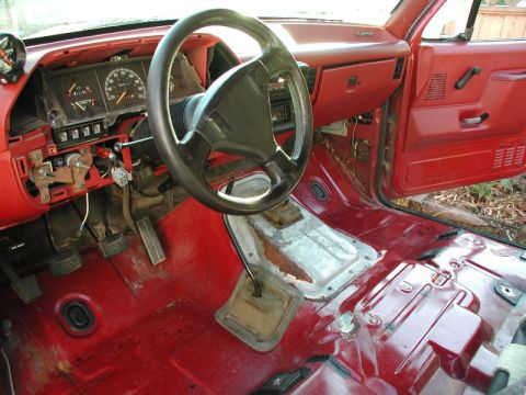 1990 Ford F-250 - The interior of a red 1990 Ford F-250 is shown, the steering wheel, dashboard, and exposed floor panels.